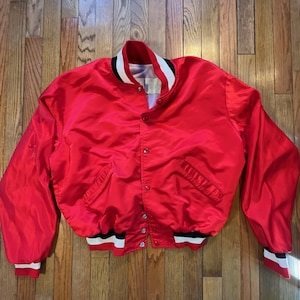 May include: A bright red satin bomber jacket with a white, black, and red striped collar, cuffs, and hem. The jacket has a button-up front, two front pockets, and a classic bomber silhouette.