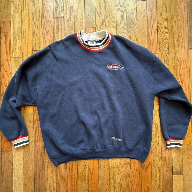 May include: Navy blue sweatshirt with the text "Field of Dreams" embroidered on the chest. The collar and cuffs have striped detailing in red, white, and navy. A vintage-style crew neck sweater.