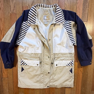 May include: Tan, navy, and white jacket with a collared neckline and multiple pockets. The jacket features a striped pattern on the collar and upper sleeves. The jacket has a drawstring waist and button closures. The jacket has the logo "S" on the chest.