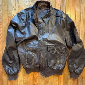 May include: A brown leather bomber jacket with a ribbed collar and cuffs. The jacket has two front pockets and a zipper closure. The brand name "DEKALB" is visible on the chest. The jacket is displayed on a wooden surface.