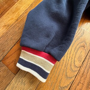 May include: Close-up of a navy blue sweatshirt sleeve. The ribbed cuff features horizontal stripes in red, tan, and navy blue. The sleeve is made of a soft, textured fabric, and the background is a wooden surface.