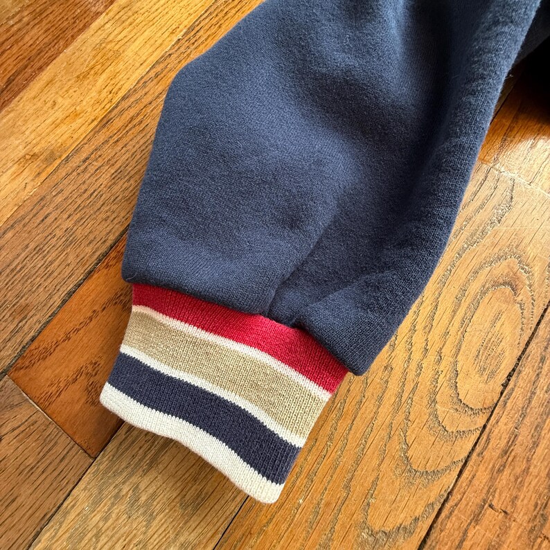 May include: Close-up of a navy blue sweatshirt sleeve. The ribbed cuff features horizontal stripes in red, tan, and navy blue. The sleeve is made of a soft, textured fabric, and the background is a wooden surface.