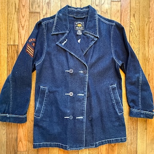 May include: A dark blue denim jacket with a double-breasted design. The jacket features a notched collar, four dark buttons, and two front pockets. An orange and gold patch is on the left sleeve. The jacket has the Route 66 logo on the label.
