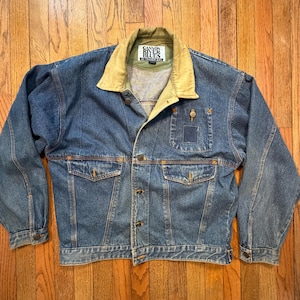 May include: A blue denim jacket with a tan collar and a button-up front. The jacket has two front pockets and a small pocket on the chest. The brand name "Canyon River Blues" is visible on the collar.