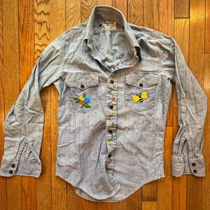 May include: Light blue button-down shirt with long sleeves. The shirt features embroidered floral and bee designs on the pockets. Colorful stitching accents the button line. The shirt has two chest pockets and buttoned cuffs.