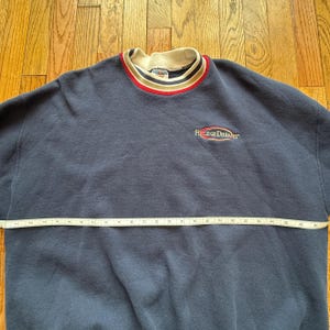 May include: Navy blue sweatshirt with a cream-colored collar and red and gold stripes. The text "Field of Dreams" is embroidered on the chest. A measuring tape is laid across the sweatshirt.