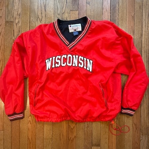 May include: A red Champion pullover windbreaker with the word "WISCONSIN" in white block letters across the chest. The jacket has a V-neck with black and white trim, matching cuffs, and a drawstring at the waist. A red whistle is attached.