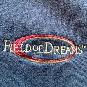 May include: Close-up of a navy blue fabric with an embroidered design. The design features the words "FIELD OF DREAMS" in a cream-colored font, enclosed within a maroon and gold oval. The fabric has a textured appearance.