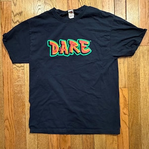 May include: A black short-sleeve t-shirt featuring the word DARE printed in a stylized graffiti font across the chest. The letters are filled with a gradient of yellow to red and outlined in a thick, vibrant green border on the dark fabric.
