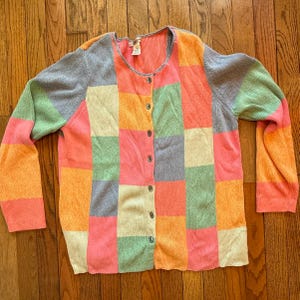 May include: A colorful patchwork cardigan sweater with a button-up front. The sweater features a variety of pastel colors, including pink, orange, green, and gray, arranged in a square pattern. The sweater is made of a ribbed knit material.