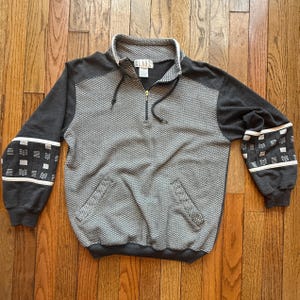May include: A gray and charcoal-colored pullover sweatshirt with a quarter-zip neckline. The body features a textured pattern, while the sleeves are solid charcoal with white and gray geometric designs. The collar is white, and the zipper is gold.