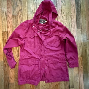 May include: A magenta-colored hooded jacket with multiple pockets and a button-up front. The jacket has a drawstring hood and a label that reads "HEFLERS". The jacket is made of a lightweight material.
