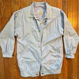 May include: A light blue jacket with long sleeves, a collar, and two front pockets. The jacket has a button-up closure and white stitching details. The collar features a striped design. The jacket is made of a lightweight material.