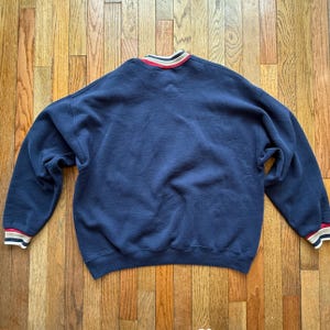 May include: Navy blue sweatshirt with a crew neck and long sleeves. The collar and cuffs feature red, white, and tan stripes. The sweatshirt is lying flat on a wooden surface.