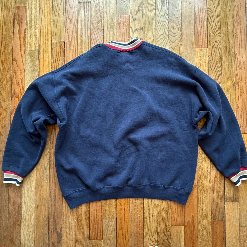 May include: Navy blue sweatshirt with a crew neck and long sleeves. The collar and cuffs feature red, white, and tan stripes. The sweatshirt is lying flat on a wooden surface.