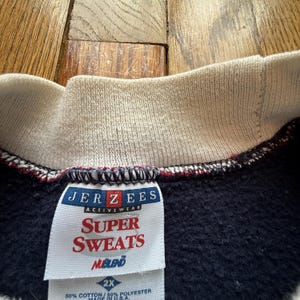 May include: Close-up of a navy blue sweatshirt with a cream-colored collar and the tag of the brand "JERZEES ACTIVEWEAR". The tag reads "SUPER SWEATS NUBLEND" and "2X". The sweatshirt is made of 50% cotton and 50% polyester.