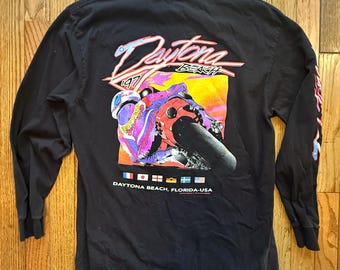 May include: Black long-sleeve shirt featuring a colorful graphic of a motorcycle racer on the back, with the words "Daytona Beach 1997". The sleeves have a flame design. The shirt also has the text "Daytona Beach, Florida, USA".