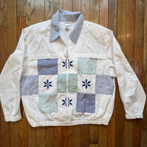 May include: White zip-up jacket with a striped collar and patchwork design. The patchwork features blue and green squares with embroidered flower details. The jacket has long sleeves and a front zipper. The brand name "ALFRED DUNNER" is visible on the collar.