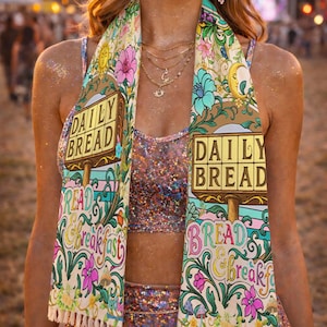 Puede incluir: Bufanda colorida con el texto "DAILY BREAD" y "BREAKFAST" en un letrero, rodeada de diseños florales y botánicos. La bufanda tiene flecos color crema y se usa sobre un top y falda brillantes. El tema general es bohemio e inspirado en festivales.