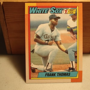 May include: A Topps baseball card featuring Frank Thomas in a light-colored uniform with the word Auburn on the chest. The card has a yellow and orange border with the text White Sox, #1 Draft Pick, and Frank Thomas printed on the front.
