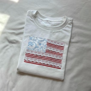 May include: White t-shirt with a unique American flag design. The flag features a blue floral print in the upper left corner and red and white striped lace detailing. The shirt is folded and rests on a white surface.