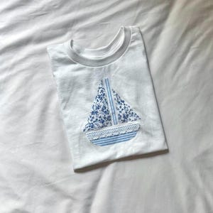 May include: White t-shirt with a sailboat applique. The sailboat features a blue floral print sail, a striped blue base, and a white lace trim. The shirt is folded on a white surface.