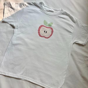 May include: White short-sleeved t-shirt with an appliqued apple design. The apple features a red and white gingham border, a light-colored center, two small black seeds, and two green leaves at the top. The shirt is made of a soft, comfortable material.
