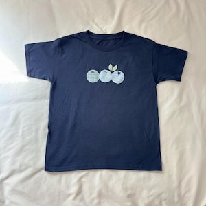 May include: Navy blue t-shirt featuring an embroidered design of three blueberries with green leaves. The blueberries have different patterns, including gingham and striped designs. The shirt is made of a soft, comfortable material.