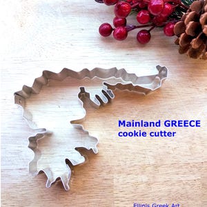 Pode incluir: Cortador de biscoitos prateado em forma da Grécia continental. O texto "Mainland GREECE cookie cutter" é visível. O cortador está sobre uma superfície de madeira, com bagas vermelhas e uma pinha no fundo.