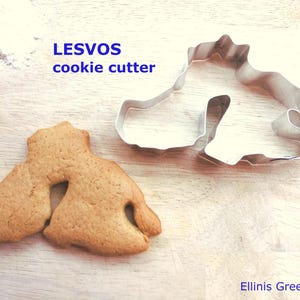 May include: A cookie cutter and a baked cookie in the shape of the Greek island Lesvos. The cookie cutter is made of silver metal. The baked cookie is golden brown. The text "LESVOS cookie cutter" is in the image.