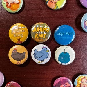 May include: A collection of 11 round buttons with various designs. The buttons feature a blue and white chicken, a blue blueberry, a black and white chicken, a yellow and red button with the text "Journey of the Prairie King", a blue button with the text "Joja Mart Life is better with Joja", a yellow and red button with the text "Junimo Kart", a purple button with a person's face, a yellow button with a person's face, a blue button with a person's face, a purple button with a person's face, and a yellow button with a person's face.
