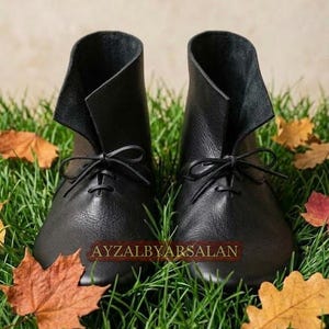 May include: A pair of black leather ankle boots with a rounded toe, a high-cut collar, and thin black laces tied in a bow at the front. The text AYZALBYARSALAN is printed in red across the center of the boots.