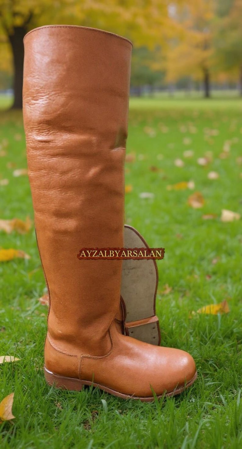 May include: A tall, knee-high brown leather boot with a rounded toe and a low heel. The boot is standing on a grassy surface, with another boot in the background. The text "AYZALBYARSALAN" is visible on the boot.