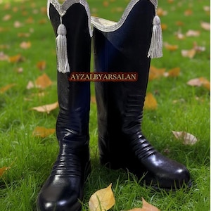 Puede incluir: Un par de botas de montar de cuero negro con adornos y borlas plateadas. Las botas tienen una caña alta y están sobre un fondo de hierba verde y hojas caídas. El texto "AYZALBYARSALAN" es visible.
