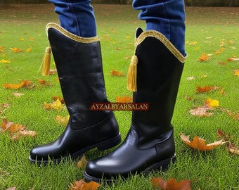 May include: Black leather knee-high boots with gold trim and tassels. The boots have a classic design and are worn with blue jeans. The text "AYZALBYARSALAN" is visible on the side of the boots. The boots are on a grassy surface.