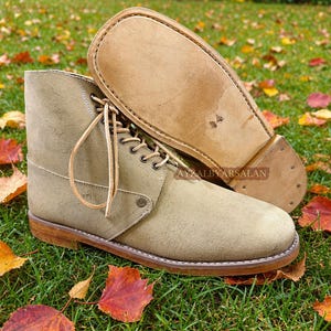 May include: A pair of beige suede ankle boots with a lace-up front, metal eyelets, and a light tan leather sole. The boots feature a rounded toe, a stacked heel, and a visible brand stamp reading AYZALBYARSALAN on the side of the sole.