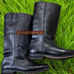 May include: A pair of black leather riding boots. The boots are tall, reaching the mid-calf, with a classic design. The boots have a smooth, polished finish and a sturdy sole. The text "AYZALBYARSALAN" is visible on the side of the boots.