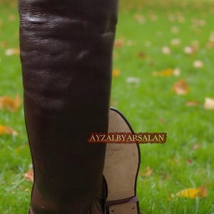 May include: A pair of tall, brown leather boots. One boot is upright, showcasing the full length, while the other lies on its side. The boots are on a grassy surface, with a blurred background of trees and foliage. The text "AYZALBYARSALAN" is visible.