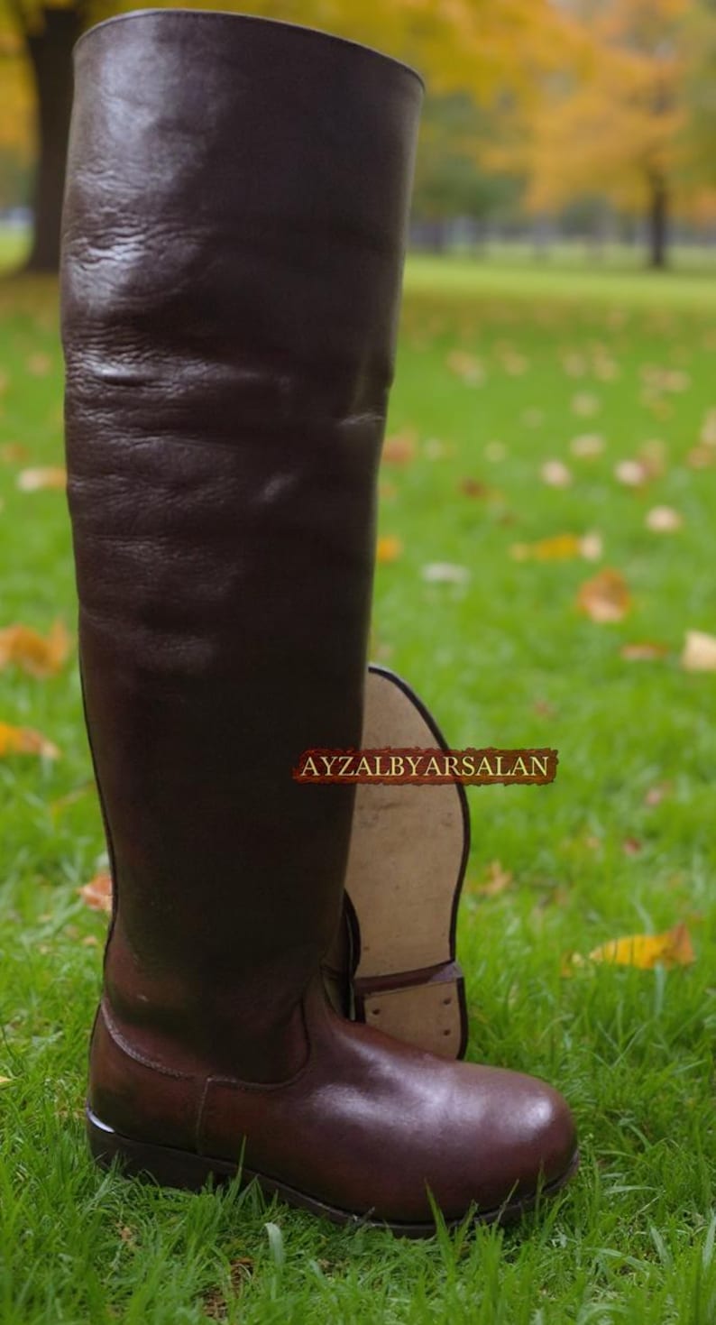 May include: A pair of tall, brown leather boots. One boot is upright, showcasing the full length, while the other lies on its side. The boots are on a grassy surface, with a blurred background of trees and foliage. The text "AYZALBYARSALAN" is visible.