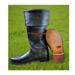 May include: Black leather boot with a folded-over top, displayed on a grassy surface. The sole of the boot is visible, showcasing its construction. The text "PAYZALBYARSALAN" is printed on the boot.