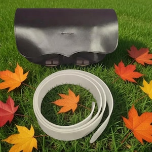 May include: A dark brown leather bag with a curved flap and two buckle closures, accompanied by a coiled light gray belt. The items are arranged on a green surface surrounded by several fallen maple leaves in shades of red, orange, and yellow.