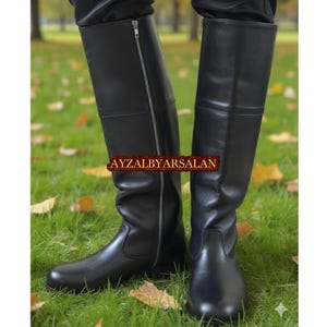 Puede incluir: Un par de botas altas de cuero negro con cremalleras laterales. Las botas son hasta la rodilla y tienen un acabado liso y pulido. El texto "AYZALBYARSALAN" es visible en la imagen.