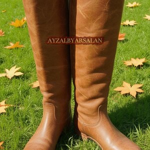 May include: A pair of tall, brown leather boots with a knee-high design. The boots are standing on green grass with fallen yellow leaves. The text "AYZALBYARSALAN" is visible on the boots.