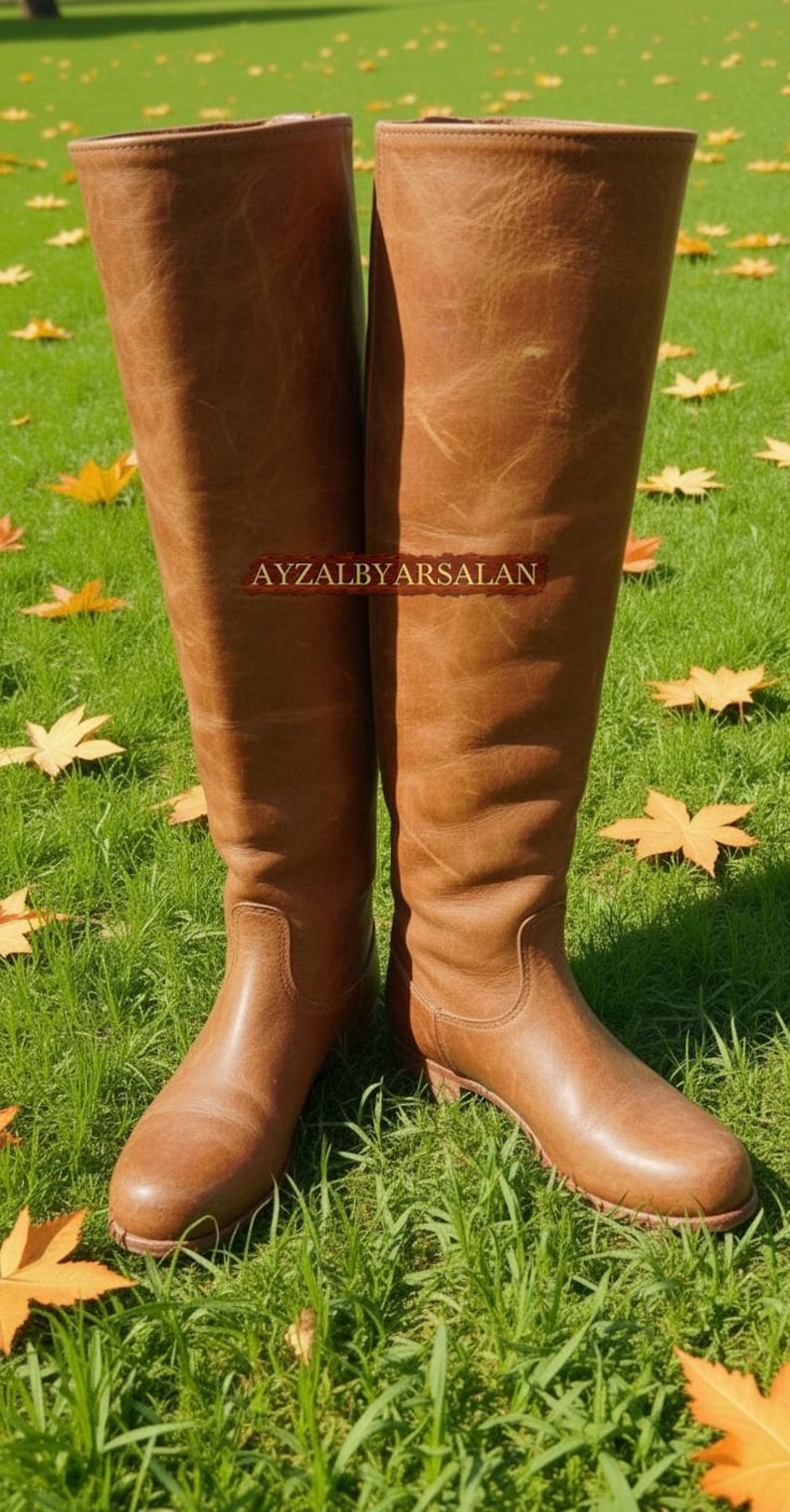 May include: A pair of tall, brown leather boots with a knee-high design. The boots are standing on green grass with fallen yellow leaves. The text "AYZALBYARSALAN" is visible on the boots.