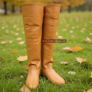 May include: A pair of tall, tan leather boots stands on a grassy lawn scattered with fallen leaves. The boots are knee-high, with a smooth texture and a slight sheen. The background is blurred, suggesting an outdoor setting.