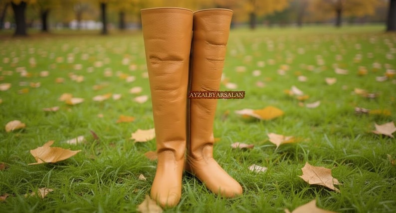 May include: A pair of tall, tan leather boots stands on a grassy lawn scattered with fallen leaves. The boots are knee-high, with a smooth texture and a slight sheen. The background is blurred, suggesting an outdoor setting.