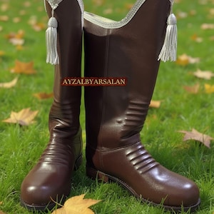 May include: A pair of brown leather riding boots with silver trim and tassels. The boots have a classic design with a smooth finish and are set against a backdrop of green grass and fallen leaves. The text "AYZALBYARSALAN" is visible.