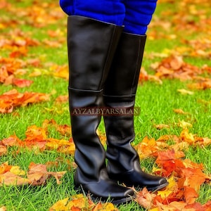 May include: A pair of black leather over-the-knee boots with a smooth finish and a rounded toe, featuring the text AYZALBYARSALAN printed across the mid-calf area. The boots are worn with bright blue trousers tucked into the top of the shafts.