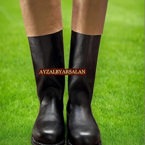 May include: A pair of black leather boots with tan-colored tops. The boots are knee-high and have a classic design. The text "AYZALBYARSALAN" is printed on the boots. The boots are set against a green background.