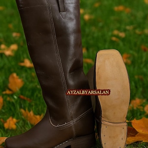Puede incluir: Un par de botas de montar de cuero marrón. Una bota está de pie, la otra está tumbada de lado, mostrando la suela. Las botas tienen un diseño clásico con un acabado liso. El texto "AYZALBYARSALAN" es visible en el lateral de las botas.